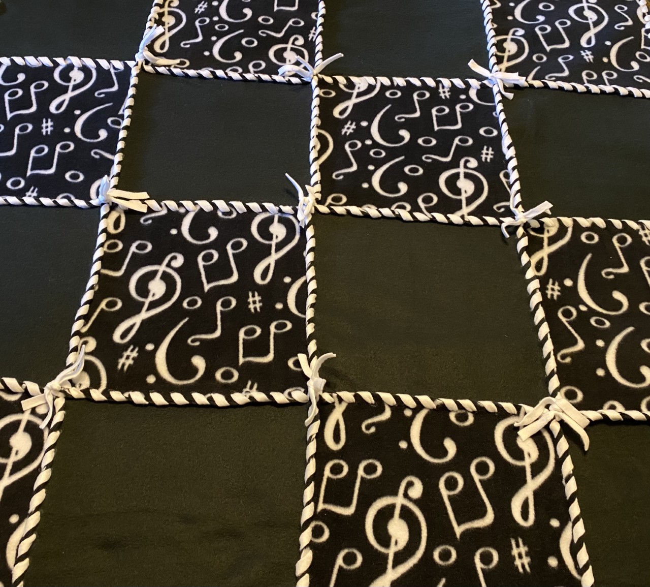 Fleece no sew 48 inch patchwork blanket with eight solid black fleece squares, alternated with eight black squares with white musical notes, woven together with white fleece ribbon