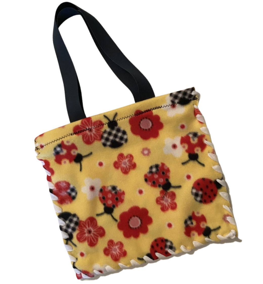 Fleece 13 inch tote bag with reinforced top edge and pre-sewn tote straps; fleece ribbon laced through tactile bordered holes around three sides, pattern red and black lady bugs, and red, white and pink floral on yellow