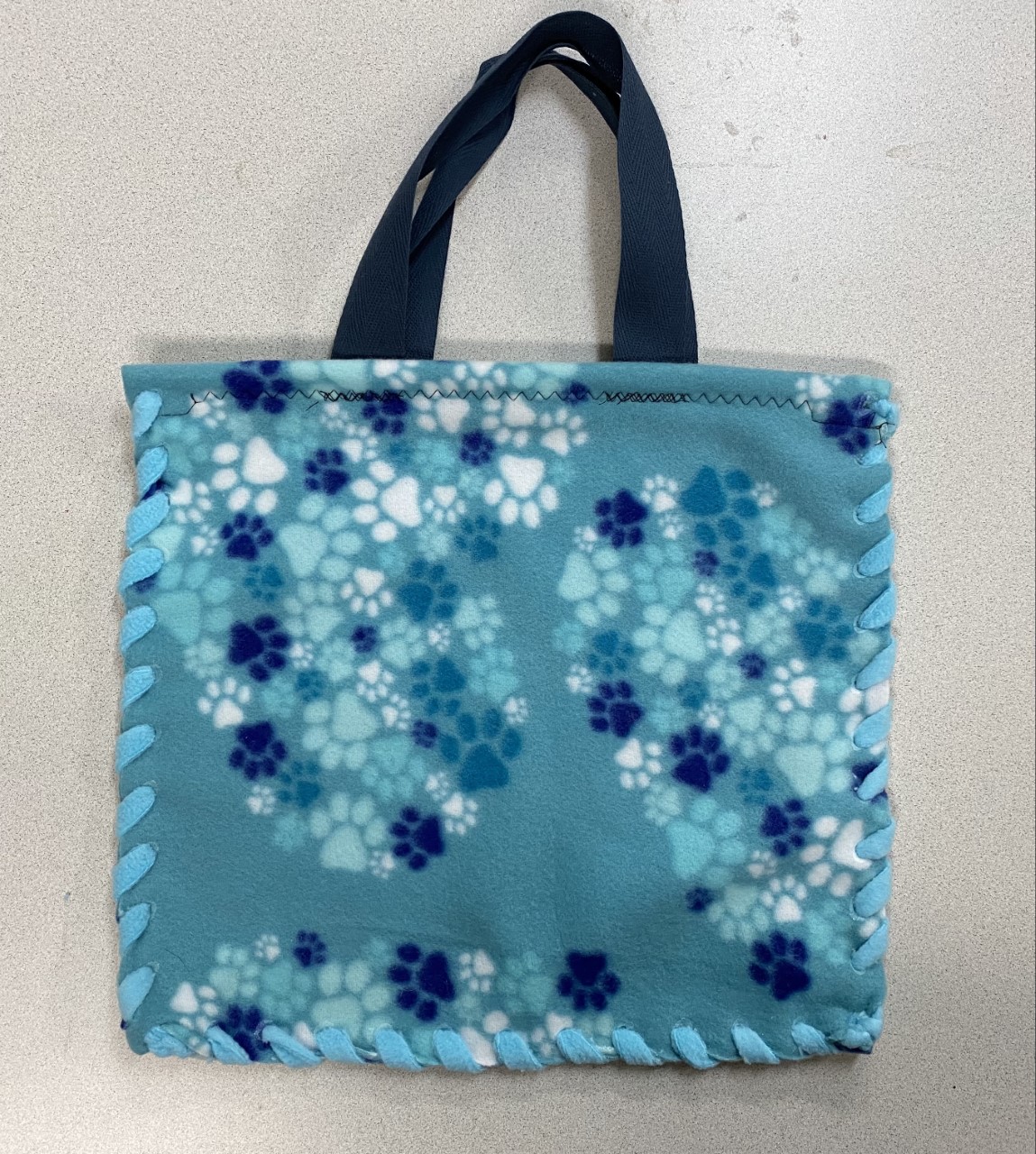 13" fleece tote bag in light teal with teal, blue and white small paw prints, blue tote straps, and teal fleece ribbon to lace with plastic needle