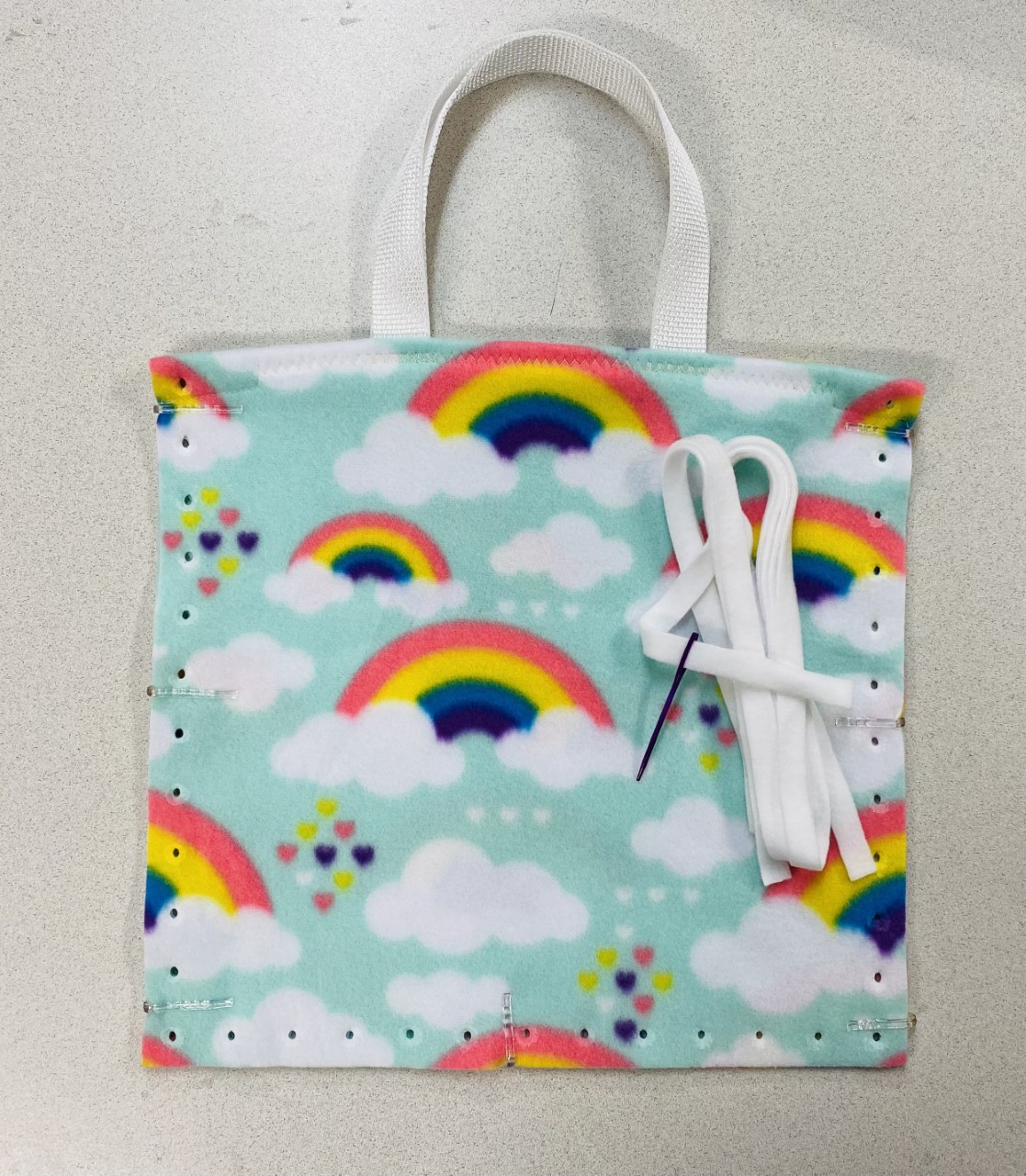 13" fleece tote bag in light teal with rainbows, clouds and small hearts, white tote straps, and white fleece ribbon to lace with plastic needle