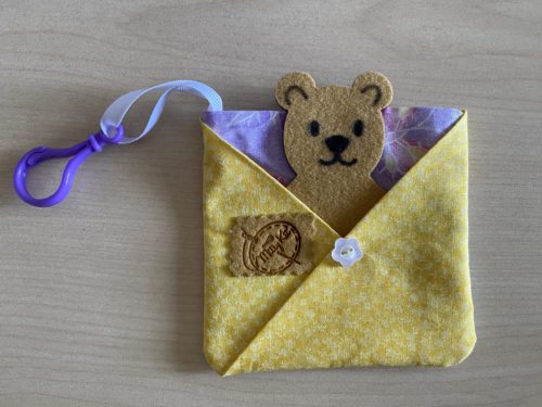 Pet in a Pocket Pouch - a light purple butterfly pocket with yellow and white floral pouch to carry a felt and faux suede teddy bear. Pouch pocket has a white floral button on center of pouch and a ribbon with hook in top left corner for attaching to loops.