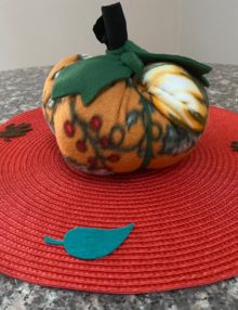 Side Photo of Fleece Pumpkin stuffed with polyfill and adorned with green leaves and black stem on top - about 8 inches around and five inches tall. The pumpkin is set on top of an orange 15" placemat decorated with felt fall leaves.