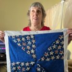 Ann Bliss holding her pinwheel quilt sewn with patriotic fabric including blue with red, white and blue stars, mottled blue, and white borders with mitered corners.